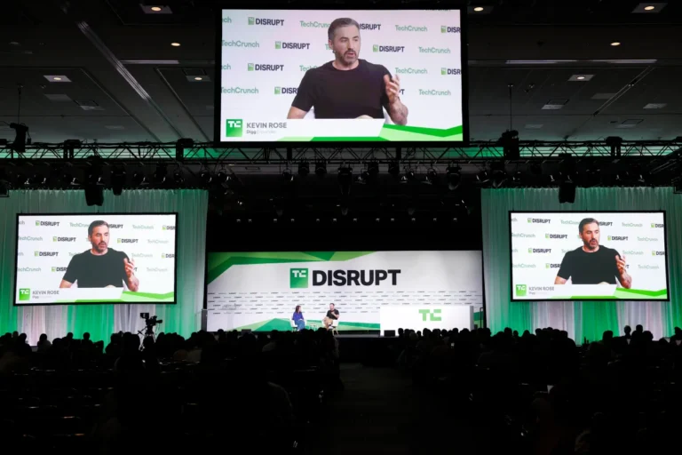 Large conference stage with "TC Disrupt" branding, speaker on screen, seated panelists, audience in foreground.