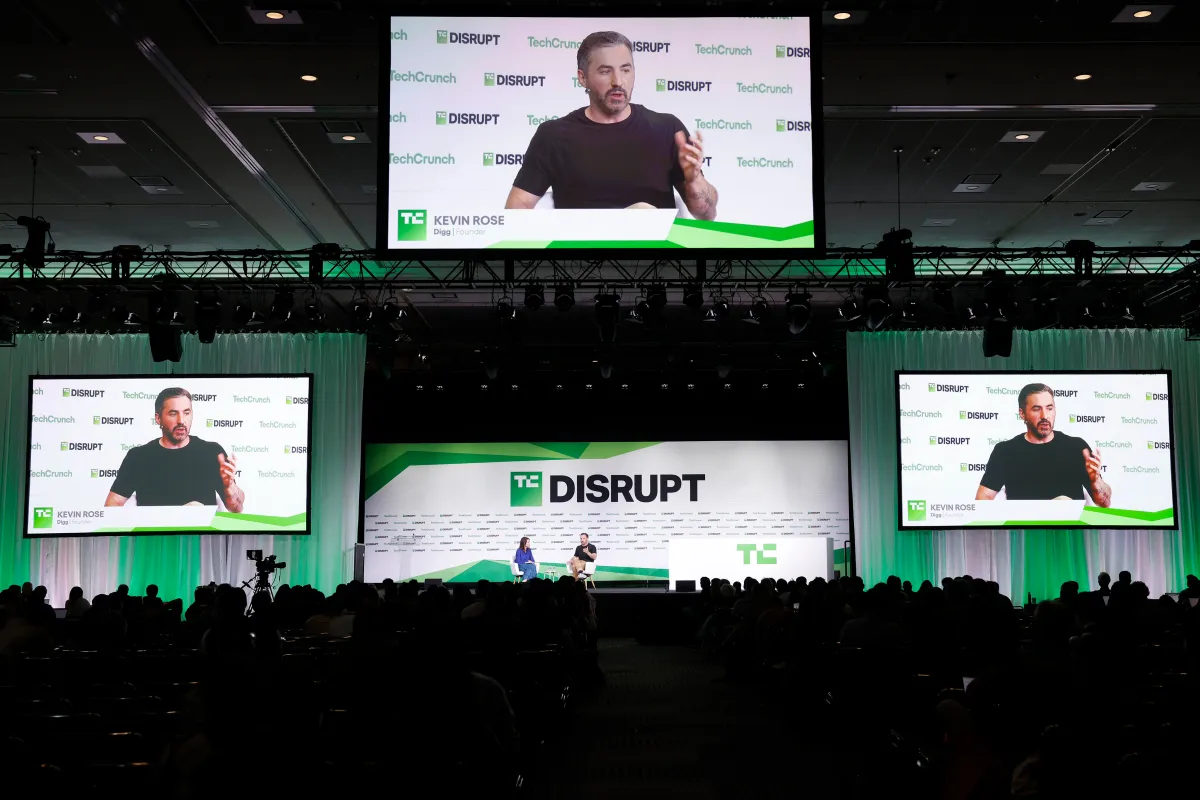 Large conference stage with "TC Disrupt" branding, speaker on screen, seated panelists, audience in foreground.