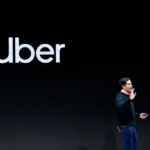 Man speaking on stage with Uber logo on black background, holding a device, gesturing with one hand.