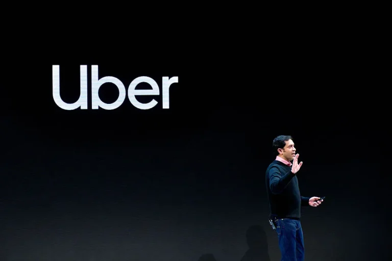 Man speaking on stage with Uber logo on black background, holding a device, gesturing with one hand.