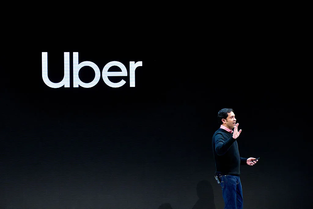 Man speaking on stage with Uber logo on black background, holding a device, gesturing with one hand.