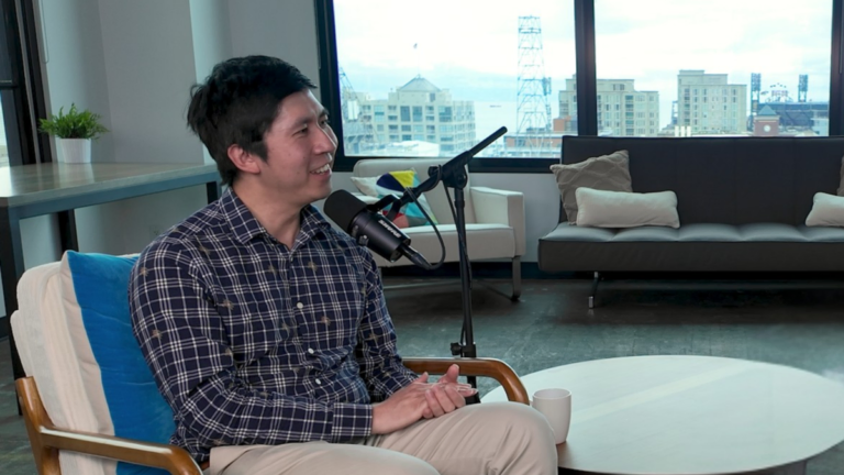 Person speaking into a microphone, seated in a modern office with large windows, couches, and a city view in the background.