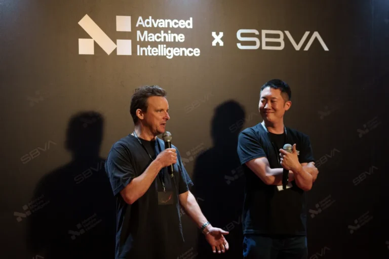 Two men on stage, one speaking into a microphone. Background text reads "Advanced Machine Intelligence x SBVA.