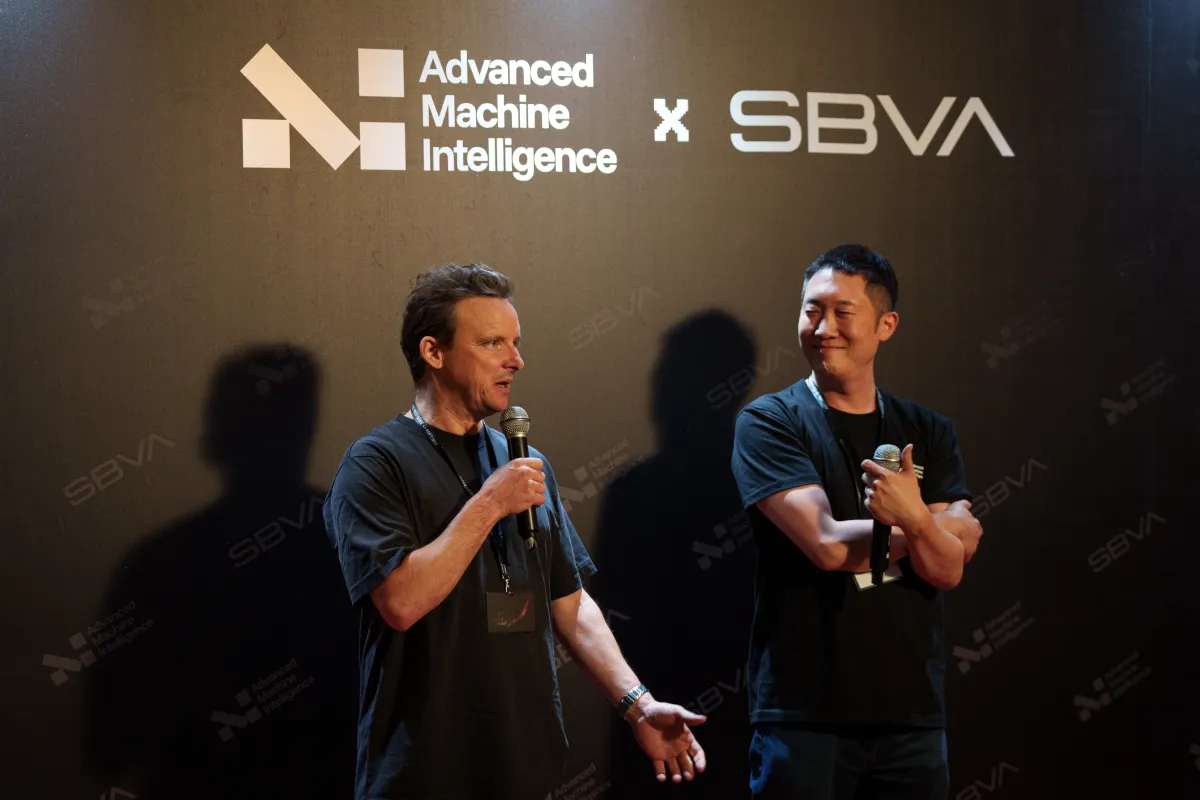 Two men on stage, one speaking into a microphone. Background text reads "Advanced Machine Intelligence x SBVA.