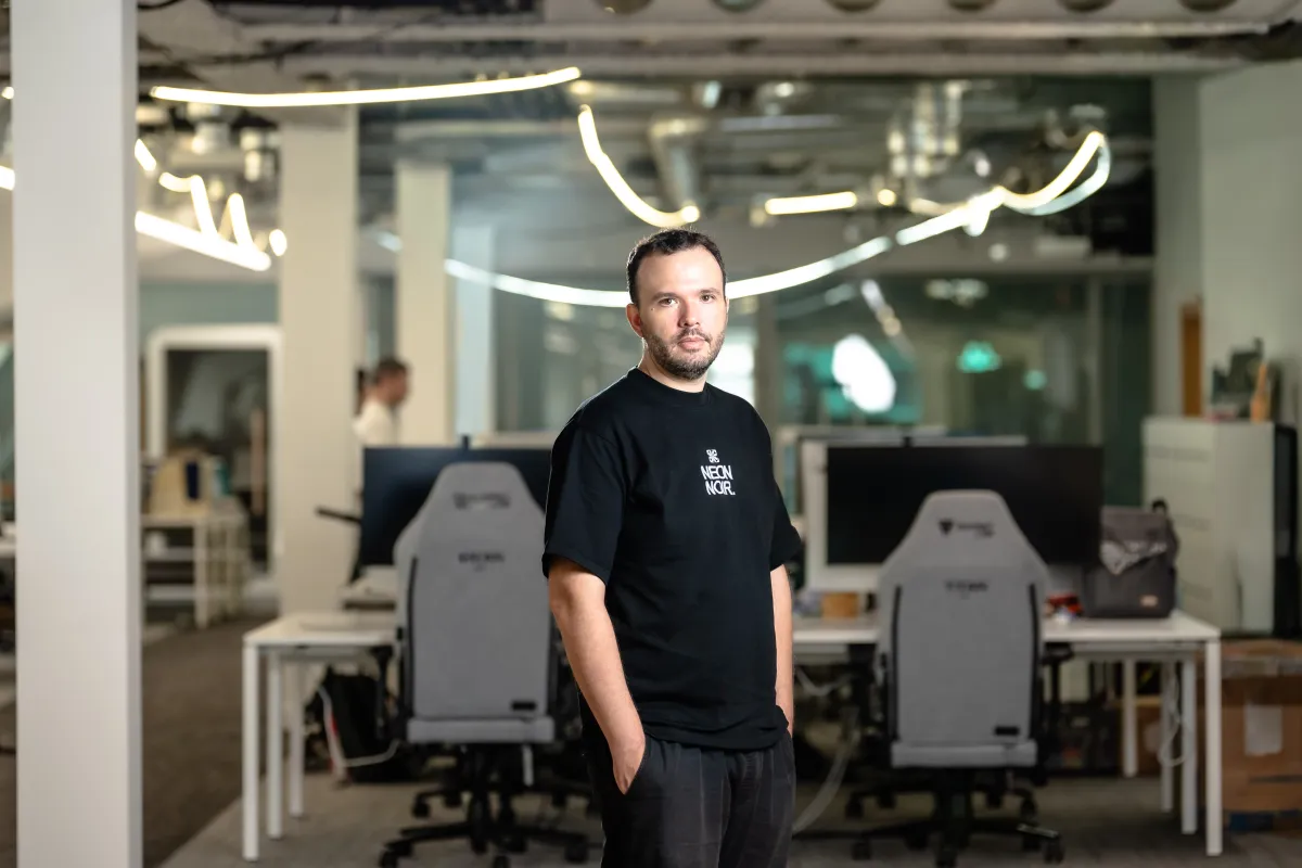 Man in black T-shirt stands in a modern office with computers and neon lights in the background.