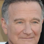 Man with short gray hair smiling slightly, wearing a dark suit, neutral background.