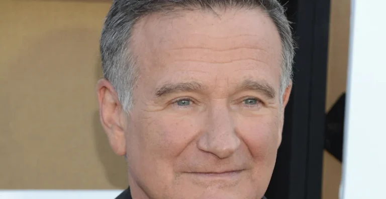 Man with short gray hair smiling slightly, wearing a dark suit, neutral background.