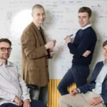Four men in an office setting, two seated and two standing, in front of a whiteboard with diagrams and notes.