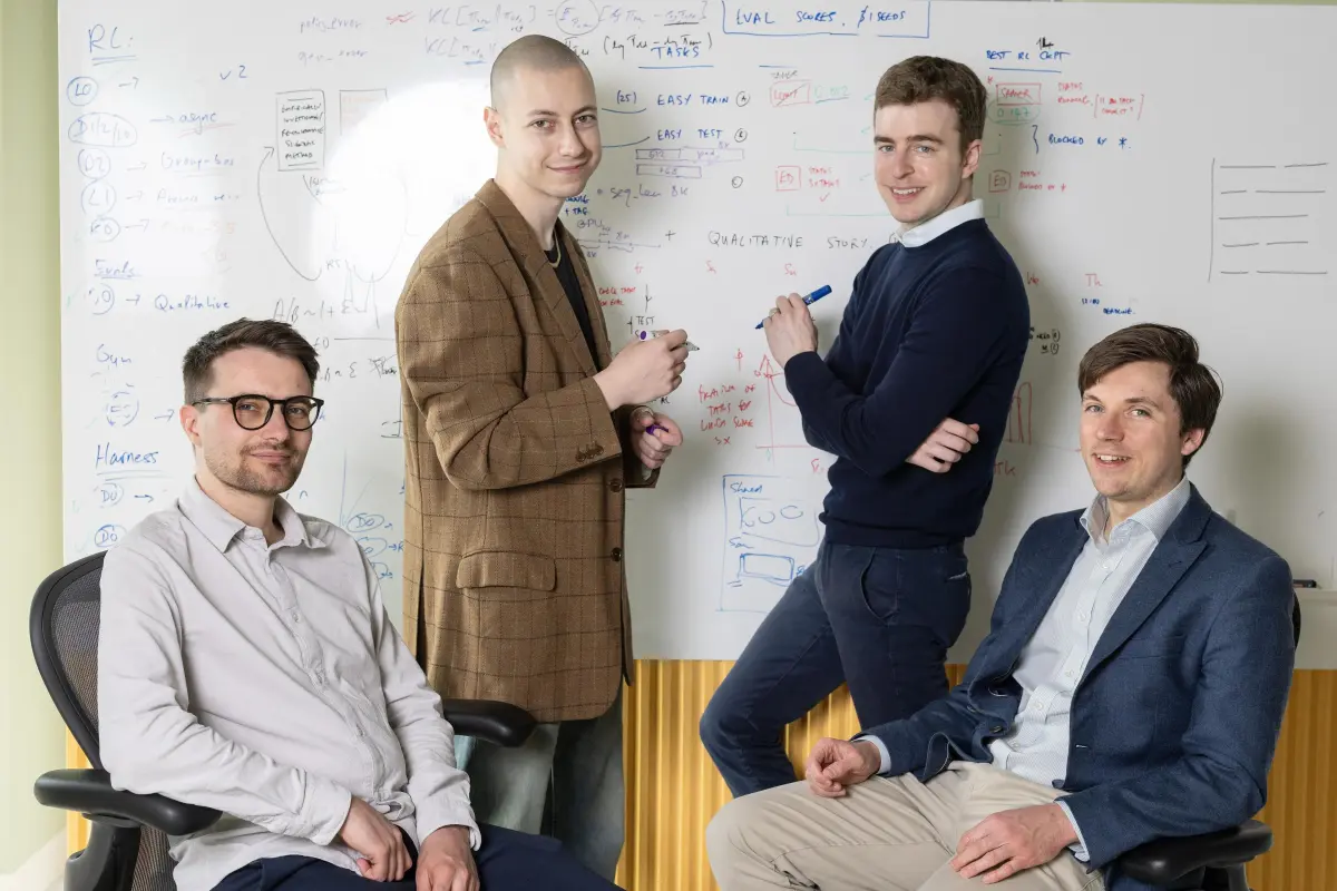Four men in an office setting, two seated and two standing, in front of a whiteboard with diagrams and notes.