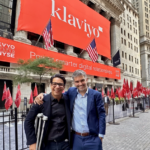 Two men smiling in front of the NYSE building with large Klaviyo banners and American flags.
