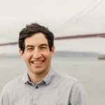 Smiling man in a gray shirt stands by water with the Golden Gate Bridge in the background on a cloudy day.