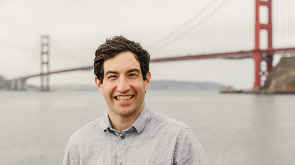 Smiling man in a gray shirt stands by water with the Golden Gate Bridge in the background on a cloudy day.