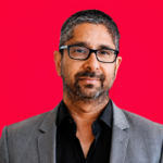 Man with glasses and beard in a gray suit and black shirt against a vibrant pink background.