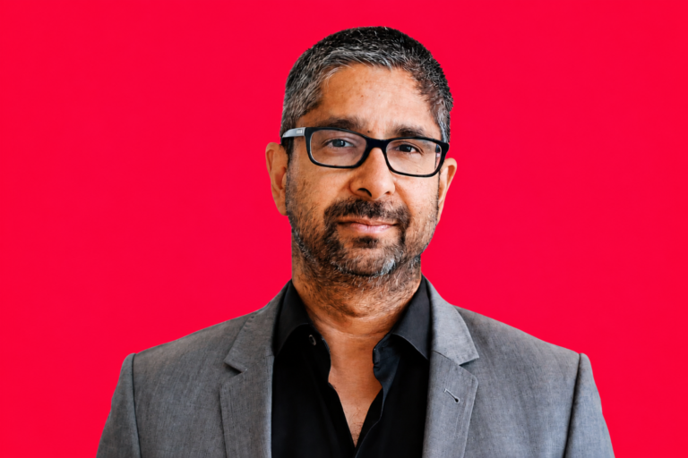 Man with glasses and beard in a gray suit and black shirt against a vibrant pink background.