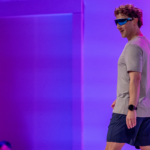 Man wearing blue-tinted sunglasses and smart watch, walking on stage with a purple-lit backdrop.