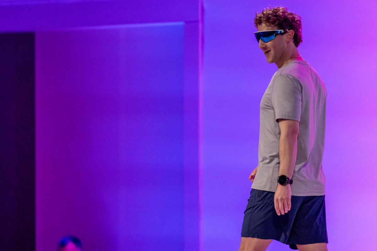 Man wearing blue-tinted sunglasses and smart watch, walking on stage with a purple-lit backdrop.