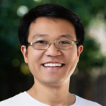 A person with glasses smiling outdoors, wearing a white shirt, with a blurred green background.