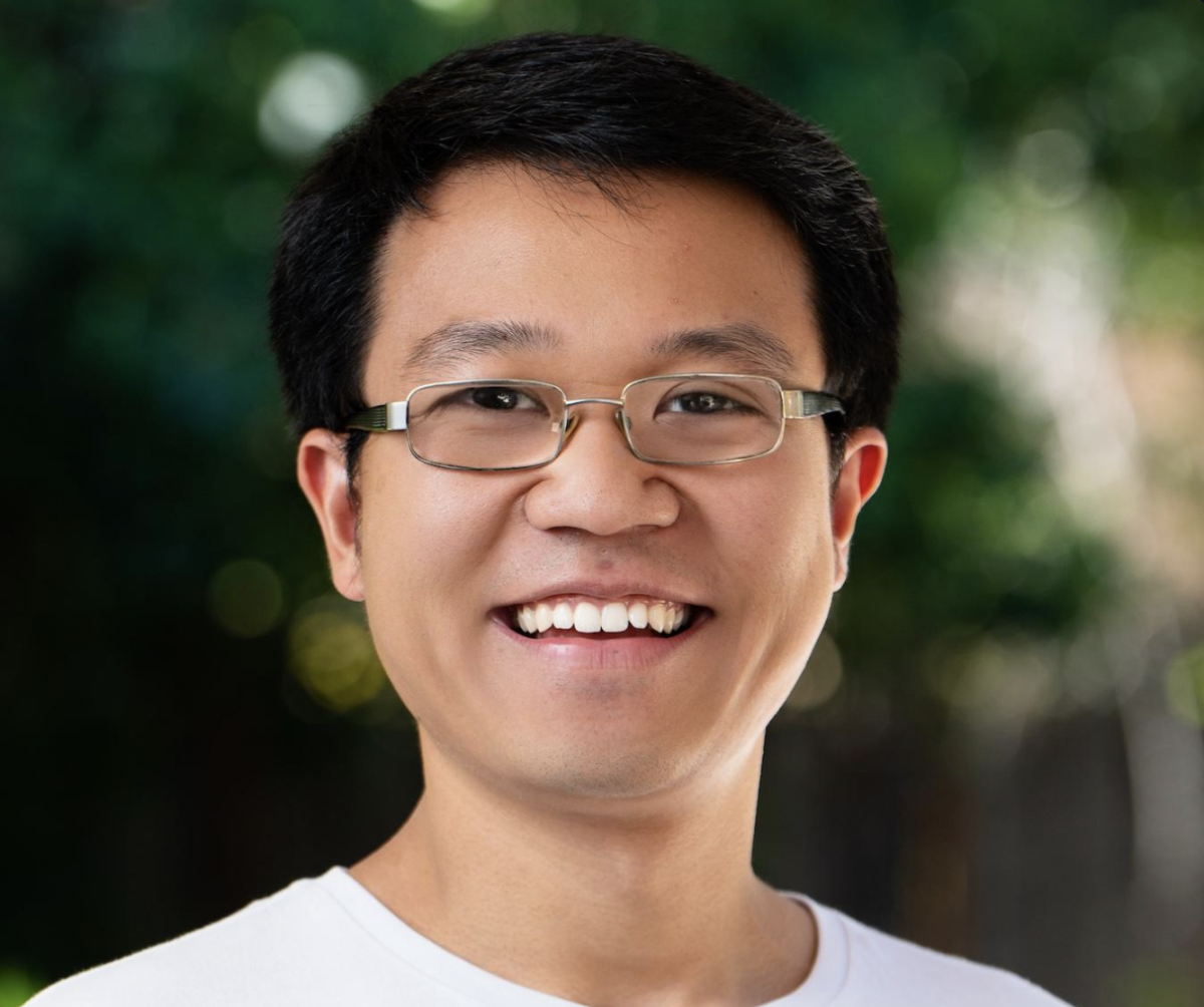 A person with glasses smiling outdoors, wearing a white shirt, with a blurred green background.