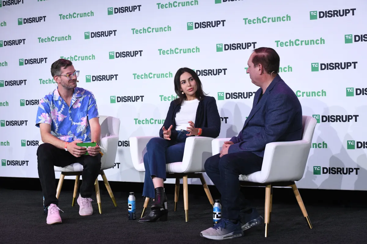 Three people seated on stage at TechCrunch Disrupt event, engaged in discussion, backdrop with event branding.