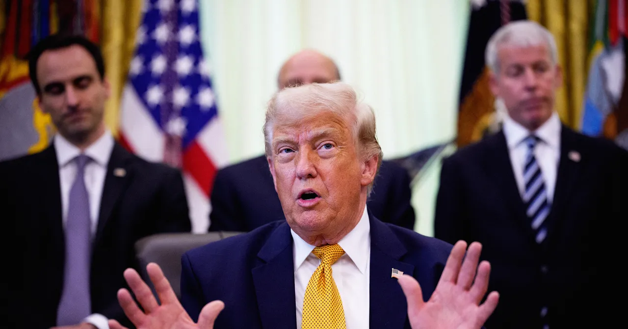 A man in a suit speaks with hands raised, three other men stand behind him in suits, flags visible in the background.