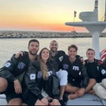 Six people smiling on a boat at sunset, wearing matching black jackets with a logo, American flag visible in the background.