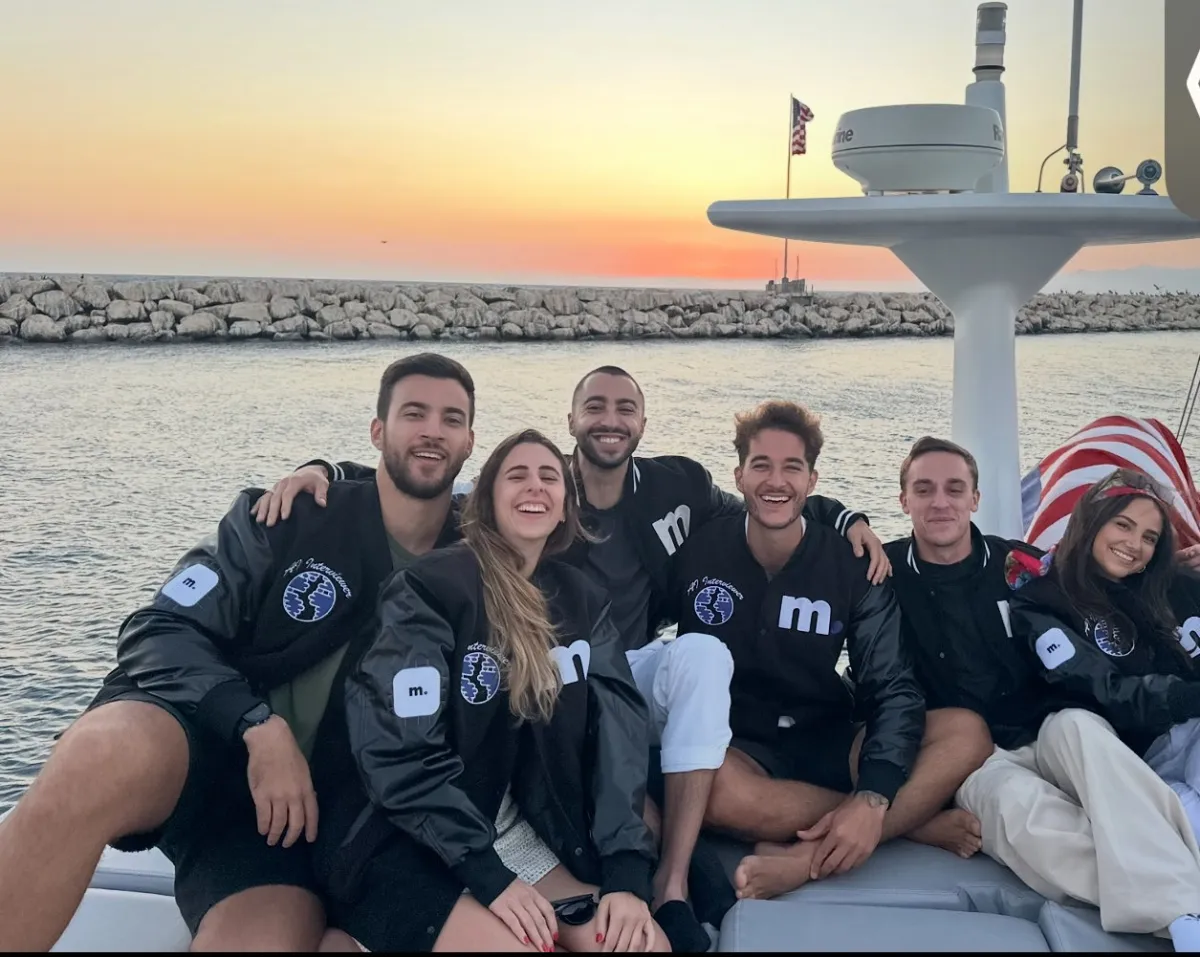 Six people smiling on a boat at sunset, wearing matching black jackets with a logo, American flag visible in the background.