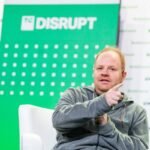 Man in gray hoodie gestures while seated, green TC Disrupt panel in background.