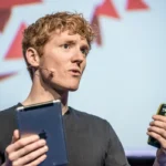 Person speaking on stage with a headset microphone, holding a tablet and a smartphone, abstract background.