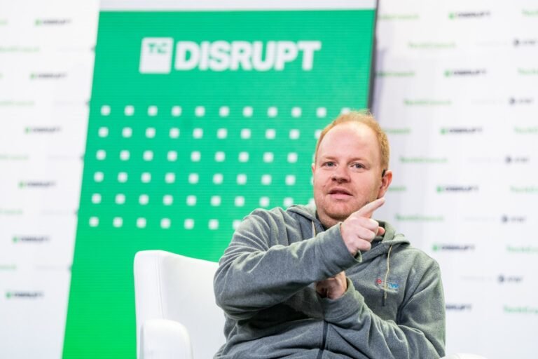 Man in gray hoodie gestures while seated, green TC Disrupt panel in background.