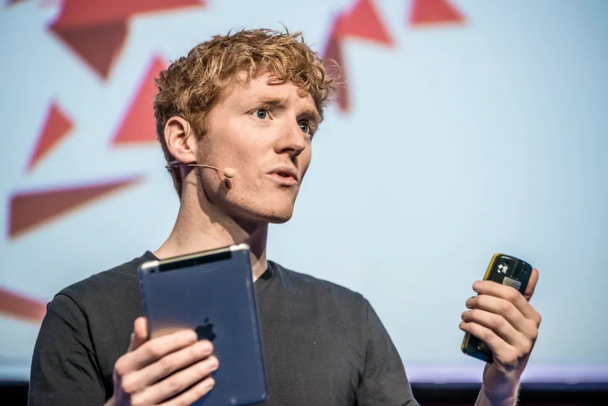 Person speaking on stage with a headset microphone, holding a tablet and a smartphone, abstract background.