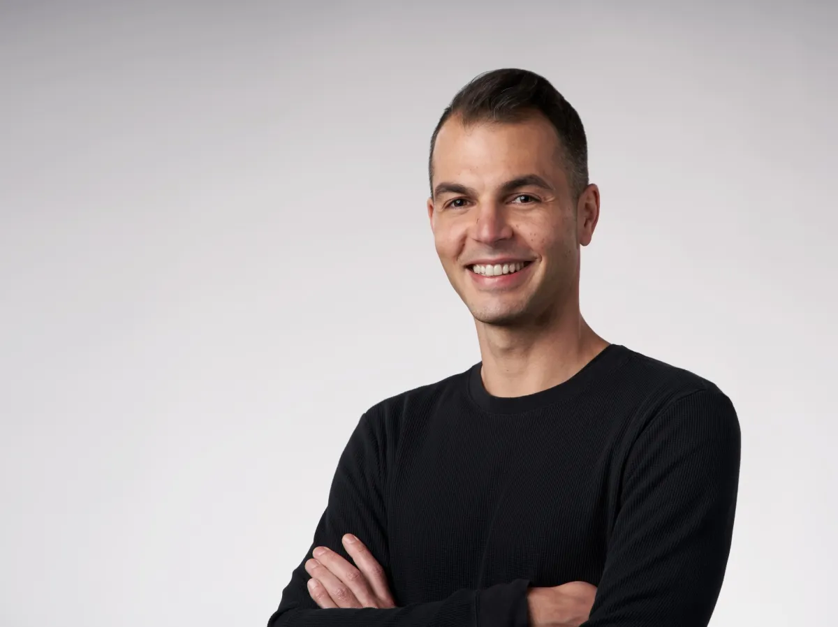 Man in a black long-sleeve shirt smiling with arms crossed, standing against a plain light gray background.
