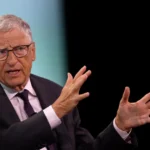 An older man in a suit and glasses gestures with both hands while speaking, against a dark background.