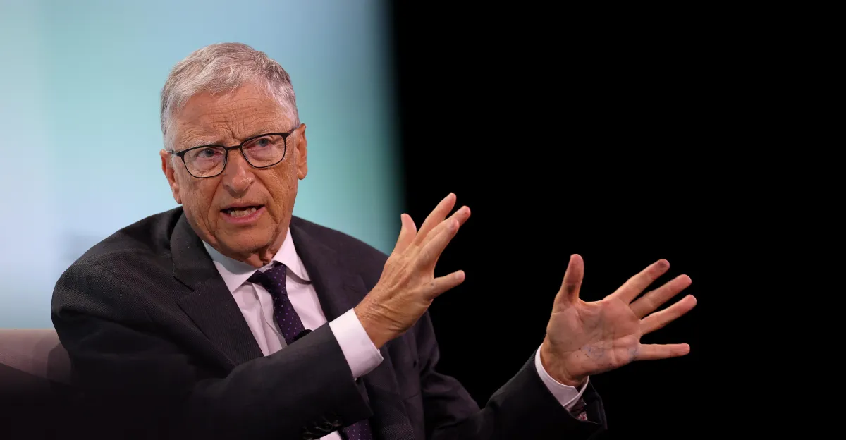 An older man in a suit and glasses gestures with both hands while speaking, against a dark background.