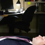 Man in a suit lying on an office floor, dimly lit by a desk lamp, surrounded by files, a computer, and office supplies.