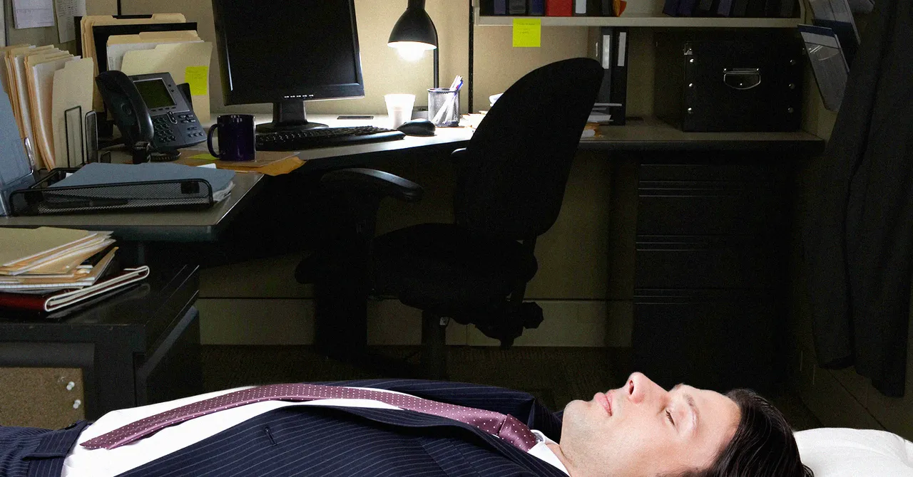 Man in a suit lying on an office floor, dimly lit by a desk lamp, surrounded by files, a computer, and office supplies.