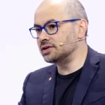 Bald man with glasses and a beard wearing a headset, speaking at an event, dressed in a dark blazer and black shirt.