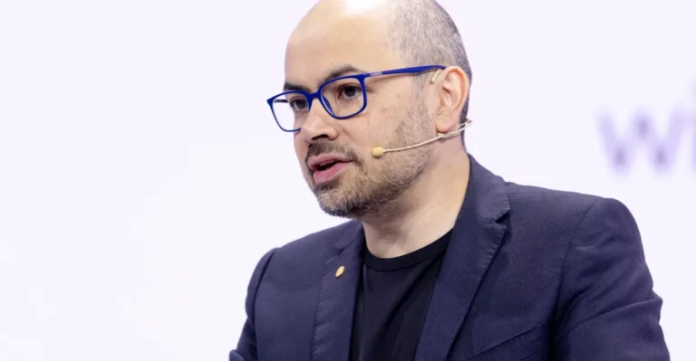 Bald man with glasses and a beard wearing a headset, speaking at an event, dressed in a dark blazer and black shirt.