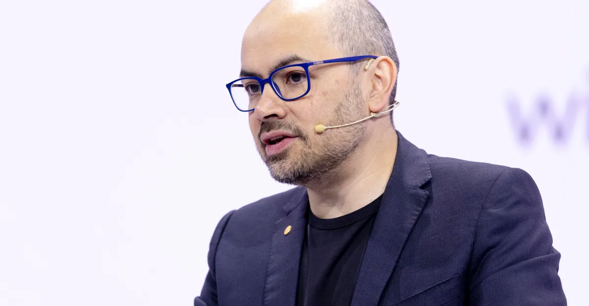 Bald man with glasses and a beard wearing a headset, speaking at an event, dressed in a dark blazer and black shirt.