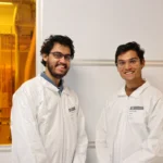 Two individuals in white lab coats and safety glasses smile in a laboratory with yellow lighting.