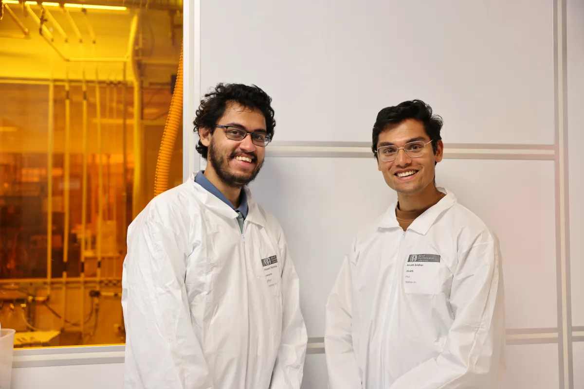 Two individuals in white lab coats and safety glasses smile in a laboratory with yellow lighting.