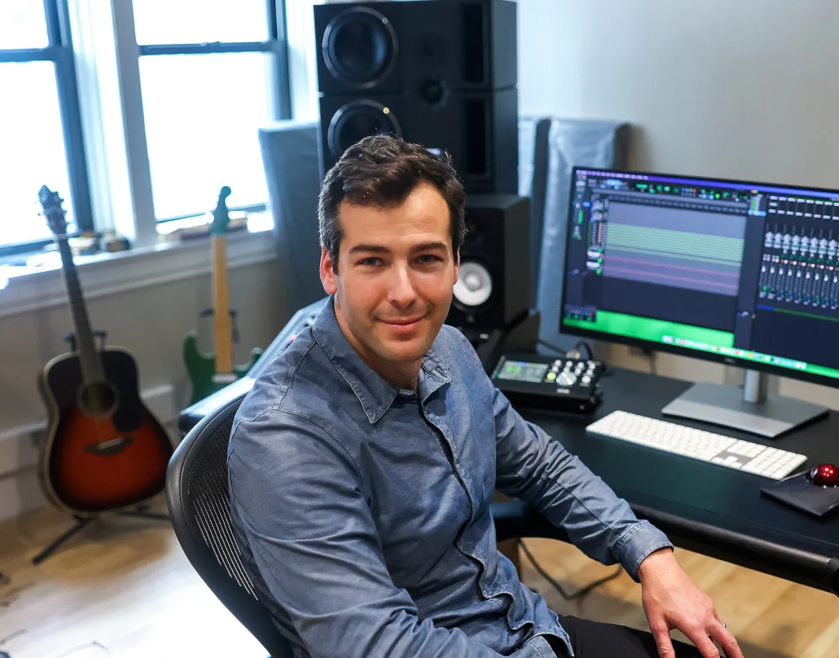Man in blue shirt seated at a desk with a computer screen displaying audio editing software, guitars in the background.