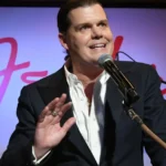 Man in a suit speaking into a microphone on stage, gesturing with his hand, with the Fender logo in the background.