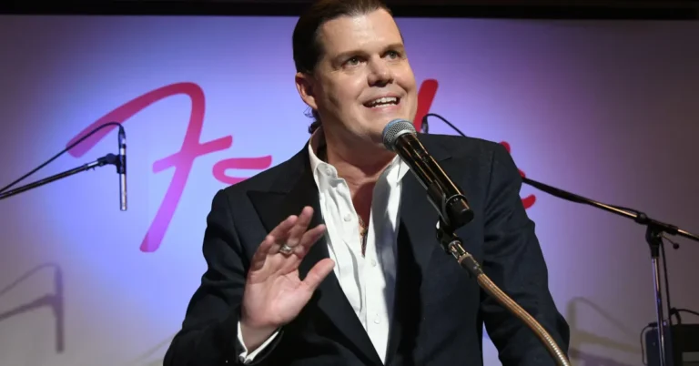 Man in a suit speaking into a microphone on stage, gesturing with his hand, with the Fender logo in the background.