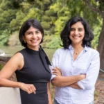 Two women smiling outdoors, one in a black dress, the other in a white shirt, with trees and a river in the background.