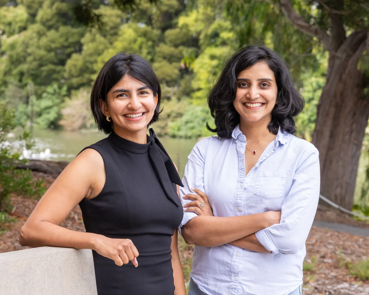 Two women smiling outdoors, one in a black dress, the other in a white shirt, with trees and a river in the background.