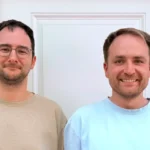 Two men standing in front of a white door, both smiling, wearing casual shirts, exterior siding visible.