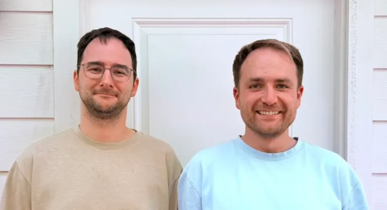 Two men standing in front of a white door, both smiling, wearing casual shirts, exterior siding visible.