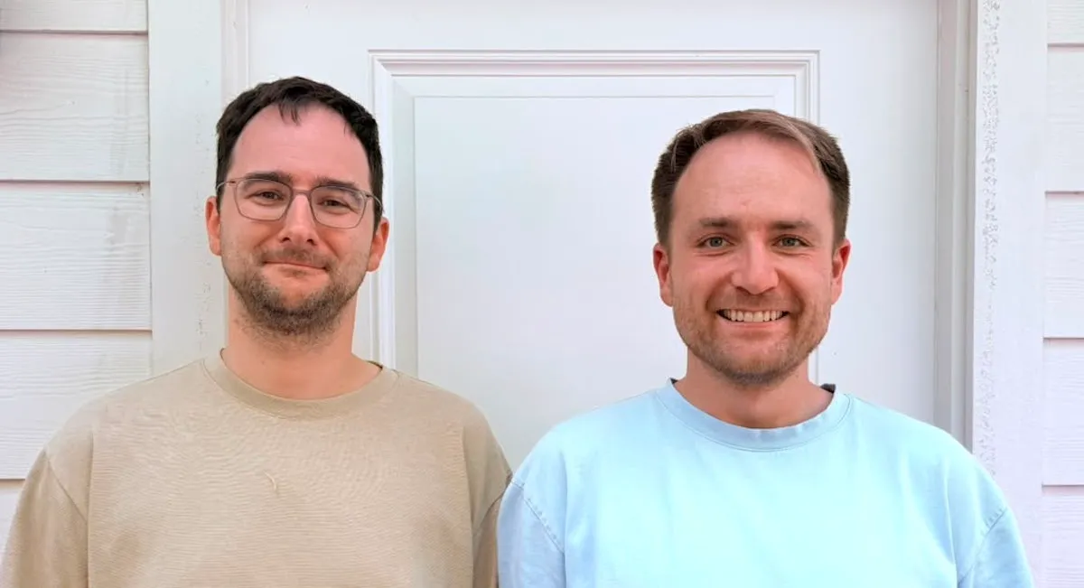 Two men standing in front of a white door, both smiling, wearing casual shirts, exterior siding visible.