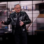 Person in a black leather jacket speaks on stage with a blurred background of a modern office interior.