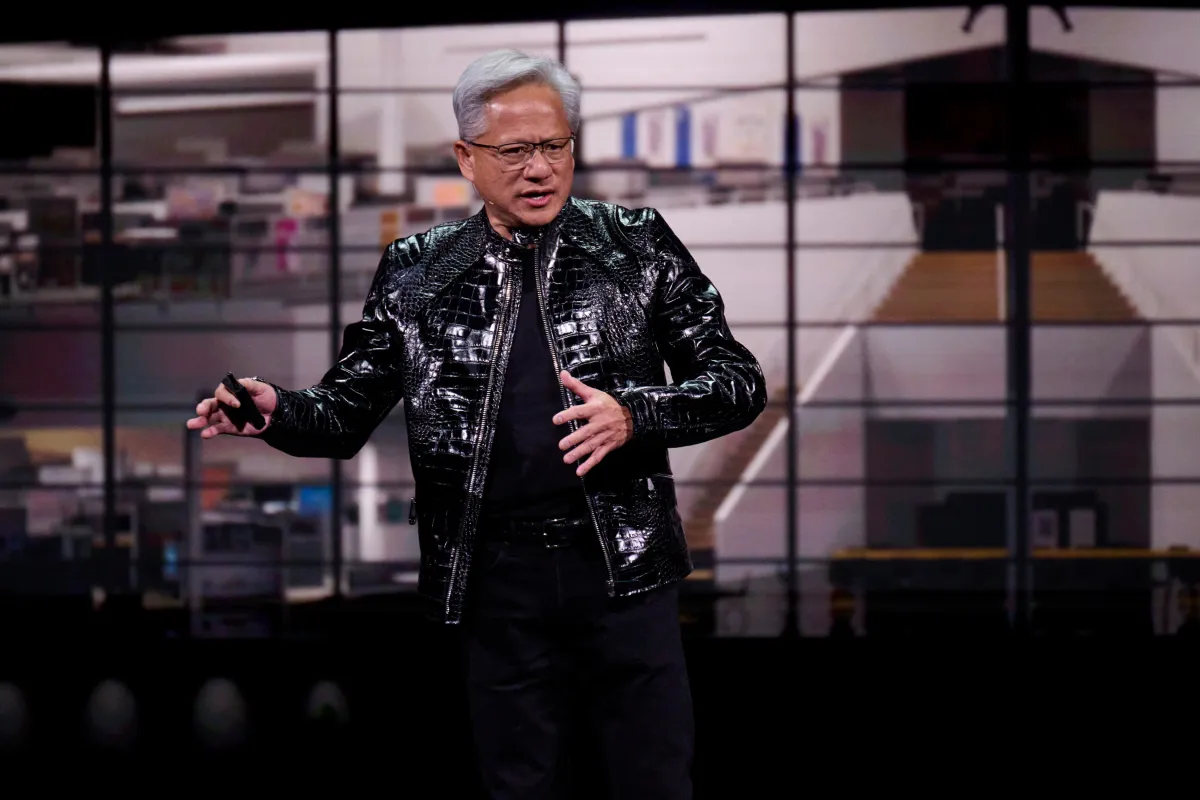 Person in a black leather jacket speaks on stage with a blurred background of a modern office interior.